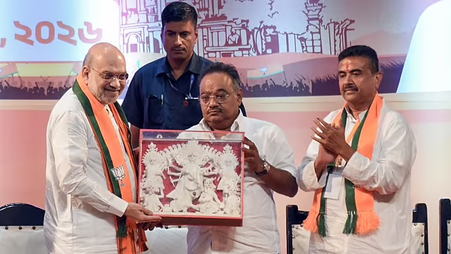 Samik Bhattacharya presenting a framed sculptural memento on stage, with Union Home Minister Amit Shah and Suvendu Adhikari alongside