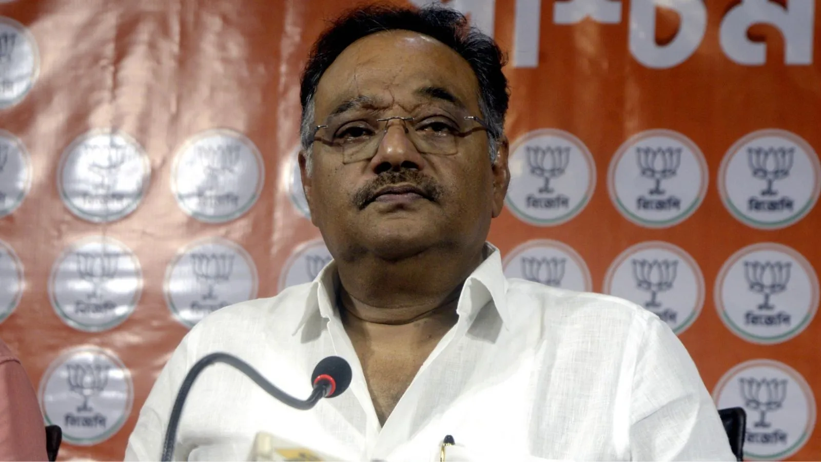 Samik Bhattacharya seated before a microphone at a party press conference, party backdrop behind him