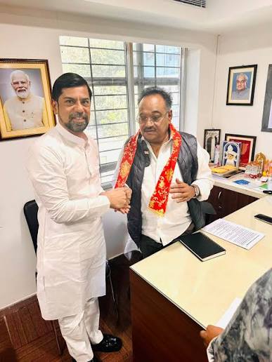 Samik Bhattacharya standing in an office shaking hands with a visitor, wearing a red ceremonial scarf