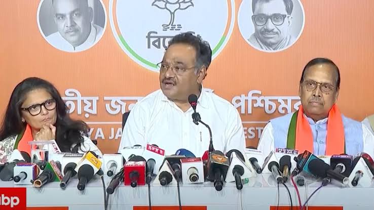 Samik Bhattacharya speaking at a press conference flanked by two colleagues, behind a dense bank of news microphones