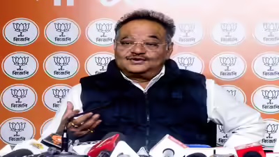 Samik Bhattacharya in a dark sleeveless jacket speaking at a press briefing in front of a wall of party emblems