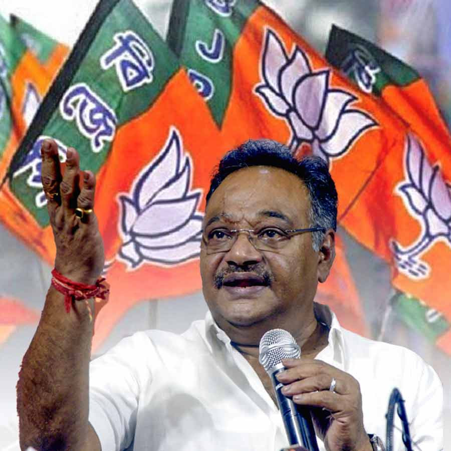 Samik Bhattacharya speaking into a microphone with his right hand raised, party flags behind him