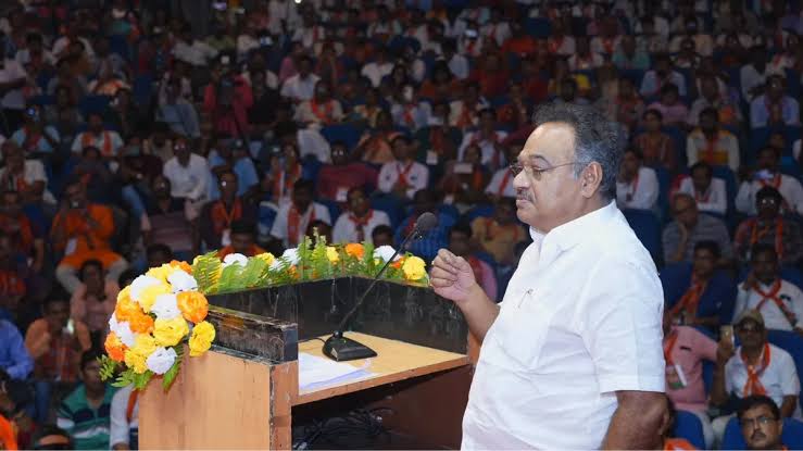 Samik Bhattacharya addressing a large seated audience from a flower-decorated lectern at an indoor convention