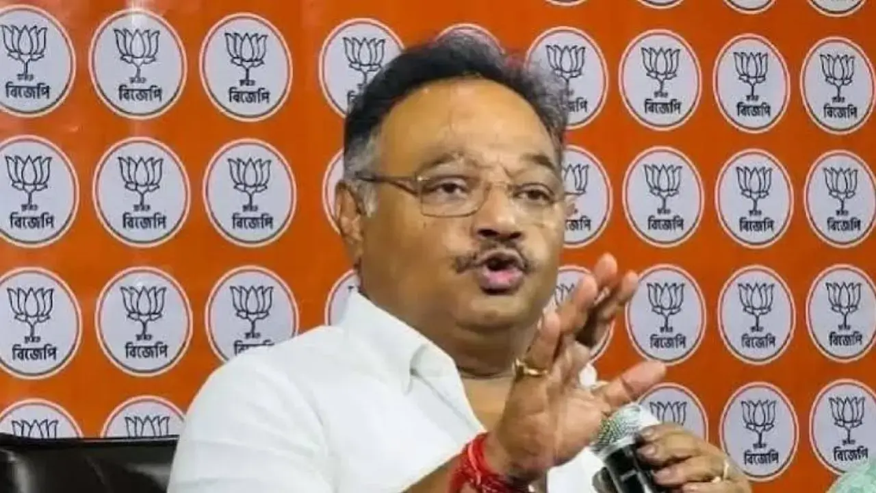 Samik Bhattacharya speaking with both hands raised in front of a party backdrop of lotus emblems