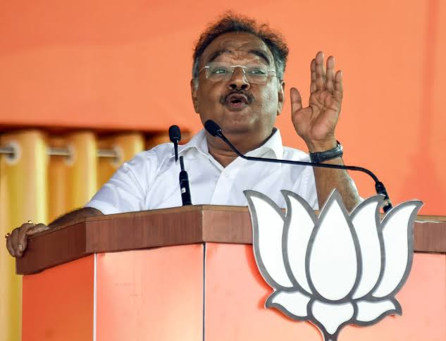 Samik Bhattacharya speaking from a podium bearing a party lotus emblem, one hand raised
