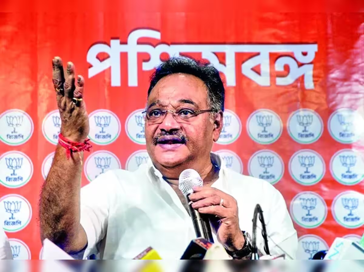 Samik Bhattacharya addressing a press briefing with a raised hand, seated before microphones with a party backdrop